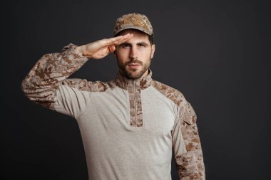 Siyah arka planda izole edilmiş kameraya bakıp selam veren kendinden emin, erkeksi bir asker.