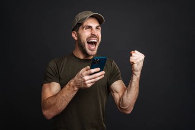 Heyecanlı asker, siyah arka planda izole bir cep telefonu kullanırken kazanan hareketi yapıyor.