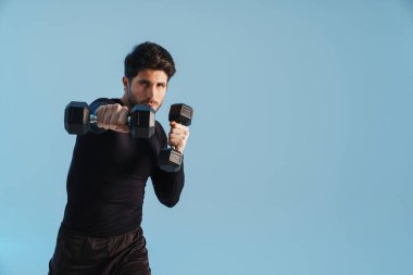 Yakışıklı sporcu mavi arka planda izole haldeki dambıllarla çalışıyor.