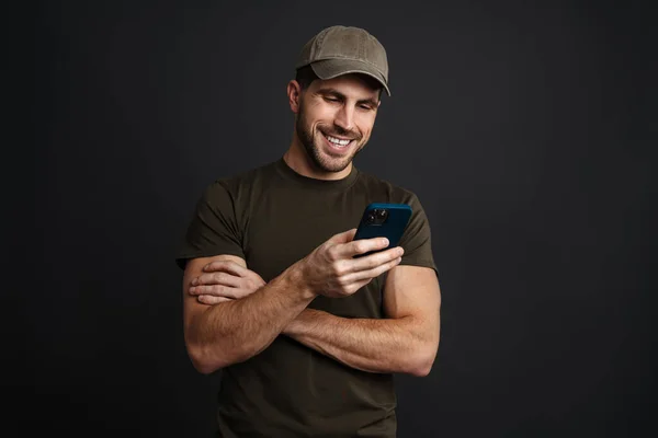 Happy masculine military man smiling and using mobile phone isolated over black background