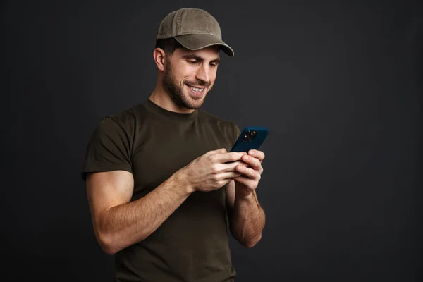 Happy masculine military man smiling and using mobile phone isolated over black background