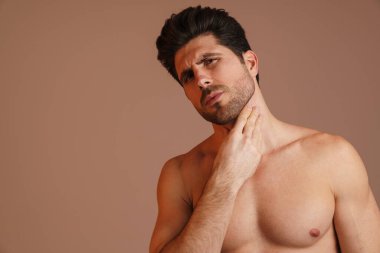 Düşüncelere dalmış yarı çıplak bir adam kameraya bakıyor ve boynu bej arka planda soyutlanmış.