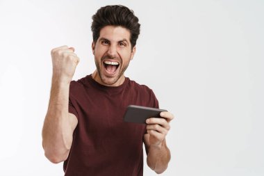 Heyecanlı adam, beyaz arka planda izole edilmiş bir cep telefonuyla çevrimiçi oyun oynarken kazanan hareketi yapıyor.