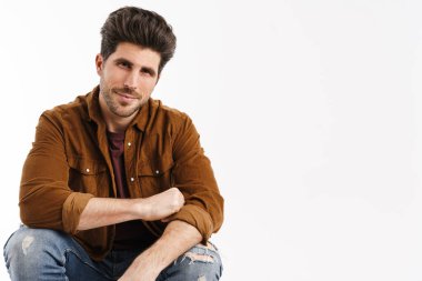 Beyaz arka planda izole bir şekilde oturan ve kameraya bakan yakışıklı genç adam.