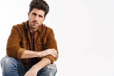 Erkeksi yakışıklı genç adam beyaz arka planda izole bir şekilde oturmuş kameraya bakıyor.