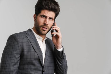 Ceketli şaşkın genç adam gri arka planda izole edilmiş bir şekilde cep telefonuyla konuşuyor ve poz veriyor.