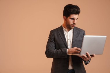 Bej arka planda dizüstü bilgisayarla çalışan ceketli ciddi bir genç adam.