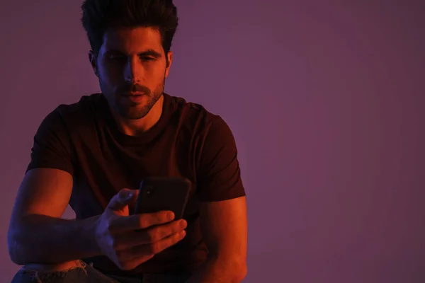 Masculine handsome man posing while using mobile phone isolated over purple background