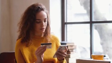 Güzel bir kadın elinde kredi kartıyla kafede otururken akıllı telefonunu kullanıyor.