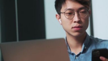 Asyalı adamın ışık ofisinde otururken telefondan laptopa bir şey kaydettiği yakın çekim görüntüsü.