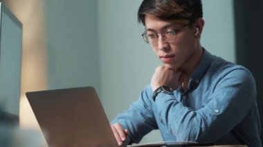 Konsantre Asyalı adam ışık ofisinde otururken dizüstü bilgisayara bakıyor.