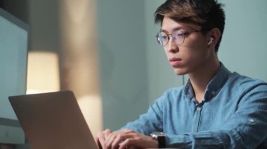 Yoğunlaştırılmış Asyalı adam dizüstü bilgisayarını karıştırıyor ve ışık ofisinde otururken ona bakıyor.