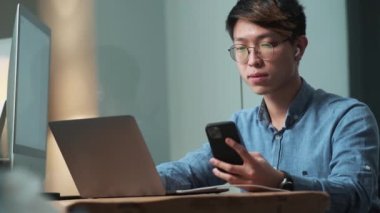 Kendine güvenen Asyalı adam ışık ofisinde otururken telefondan laptopa bir şeyler kaydediyor.