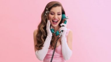 Eldivenleri ve saç tokaları ile muhteşem pembe elbiseler giyen mutlu genç bir kadın stüdyodaki pembe bir duvarda izole edilmiş eski bir telefondan konuşuyor.