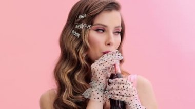 Eldivenleri ve saç tokaları ile muhteşem pembe elbiseler giyen genç bir kadın stüdyodaki pembe bir duvarın üzerinde izole bir şekilde duran cam şişeden kola içiyor.