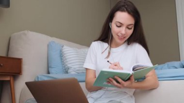 Güzel bir kadın evdeki yatak odasında otururken bilgisayarını kullanarak notlar alıyor.