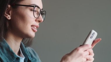 Gözlüklü, telefonla mesajlaşan ve mutfaktaki masada otururken ileriye bakan mutlu kadının yakın görüntüsü.