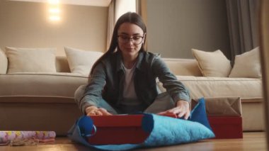 Gözlüklü gülümseyen kadın odada yerde otururken hediyesini hediye kağıdına sarıyordu.