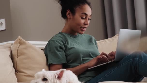 Egy Mosolygó Afro Amerikai Használja Laptopját Miközben Otthon Kanapén Kutyájával — Stock videók