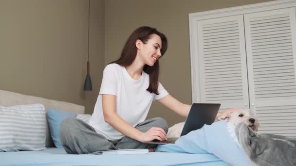 Gülümseyen Bir Kadın Evinde Köpeğiyle Yatak Odasında Otururken Bilgisayarını Kullanıyor — Stok video