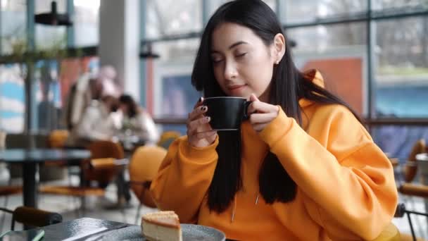 身穿橙色帽衫的微笑的女人 坐在咖啡店里 一边喝着杯子里的酒味 一边环顾四周 — 图库视频影像