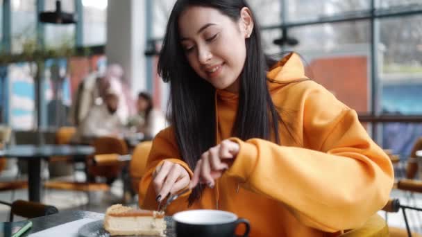 一个穿着橙色帽衫的微笑的女人坐在咖啡店里 一边切着一块蛋糕一边欣赏着它 — 图库视频影像