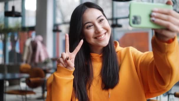 一位穿着橙色帽衫的积极女子坐在咖啡店里 一边在电话里自拍 一边伸出大拇指 摆出和平的架势 — 图库视频影像