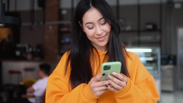 一个穿着橙色帽衫的笑女人站在咖啡店里看着电话 在电话上打字 — 图库视频影像