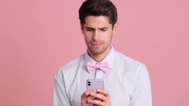 Papyon takan mutlu genç bir adam, stüdyodaki pembe bir duvarın üzerinde tek başına beklerken akıllı telefonunu kullanıyor.