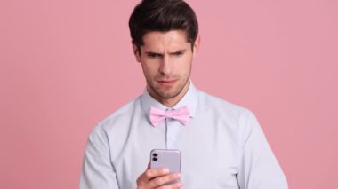 Papyon takan hoşnutsuz genç bir adam stüdyodaki pembe bir duvarın üzerinde duran akıllı telefonuna bakıyor.