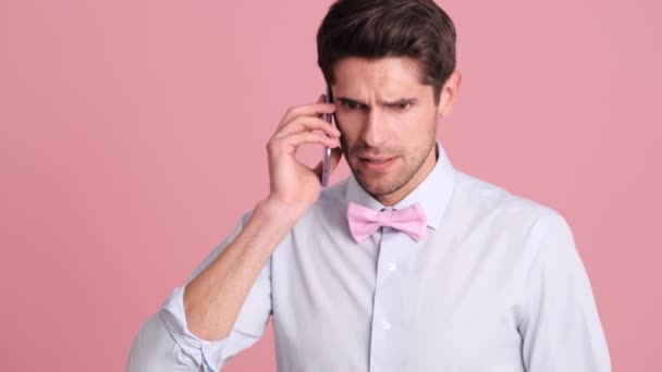 一位戴着蝴蝶结领带的震惊的年轻人站在工作室的粉红墙上 用智能手机与人交谈 — 图库视频影像