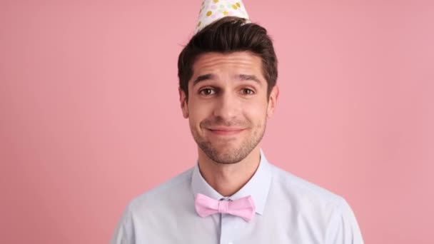 一位戴着生日礼帽 面带微笑的年轻人正与站在工作室粉红墙上的摄像机打招呼 — 图库视频影像