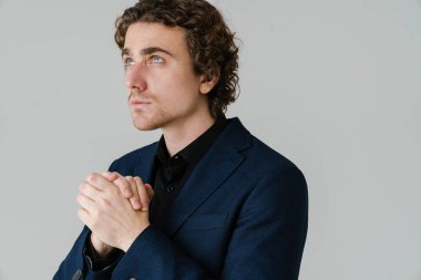 Beyaz arka planda avuç içlerini bir arada tutan kafası karışmış, ceketli yakışıklı bir adam.