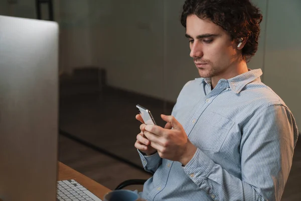 Caucasian young serious businessman in earphones using cellphone while working on computer in office