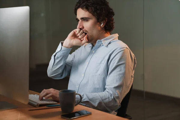 Caucasian young businessman in earphones yawning while working on computer in office