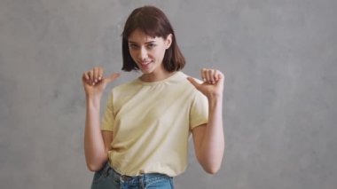 Gri stüdyoda dikilirken başparmağıyla işaret eden kısa saçlı pozitif kız.