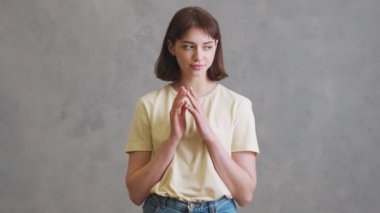 Kısa saçlı pozitif bir kız, gri stüdyoda dikilirken bir şeyler düşünüyor ve parmaklıyor.