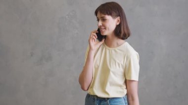 Gri stüdyoda dikilirken telefonda konuşan kısa saçlı, gülümseyen kız.