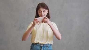 Kısa saçlı mutlu kız, gri stüdyoda dikilirken telefonda oyun oynuyor.