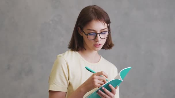 グレーのスタジオに立っている間にノートに何かを書いている眼鏡の中の大胆な女の子 — ストック動画