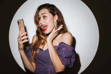 Gece elbisesi içinde, projektör ışığından izole edilmiş cep telefonu kullanan heyecanlı güzel kız.