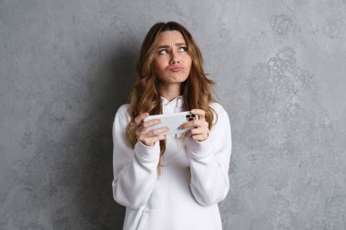 Gri arka planda izole edilmiş bir cep telefonuyla oynayan gündelik giysili düşünceli genç kız.