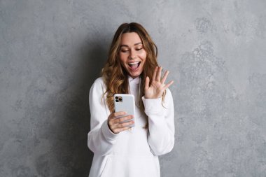 Gri arka planda izole edilmiş bir video görüşmesinde cep telefonlu neşeli, heyecanlı genç bir kadın.