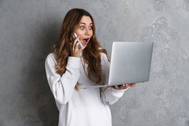 Gri arka planda tek başına dururken dizüstü bilgisayar ve cep telefonu kullanan neşeli genç ve çekici bir kadın.