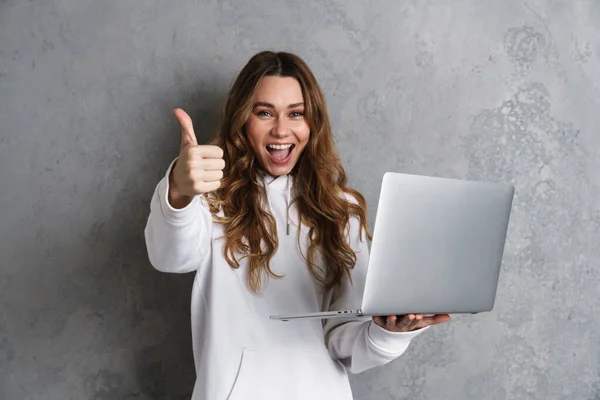 快乐而兴奋的年轻女性穿着休闲装 用笔记本电脑与灰色背景隔离 竖起大拇指 — 图库照片