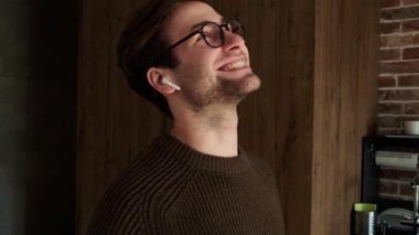 Süveter giyen, kablosuz kulaklıkla konuşan ve mutfakta dikilirken el kol hareketi yapan bir adam.