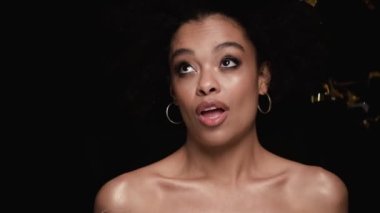 Güzel, gülümseyen bir Afrikalı Amerikalı kadın kameraya poz veriyor. Stüdyonun siyah arka planında tek başına duruyor. Altın konfetileri düşüyor.