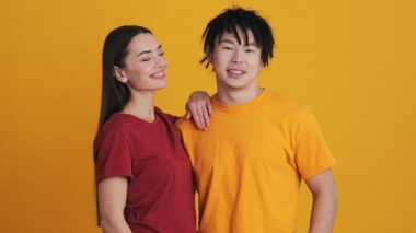 Kadın ve erkek kameraya poz verirken, gülümseyen çok uluslu çift stüdyoda sarı duvarın üzerinde erkeğini öpüyor.