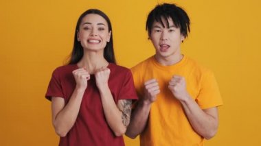 Mutlu çokuluslu çift, kazanan jest yapıyor. Stüdyonun sarı duvarında izole bir şekilde duran kameraya bakıyor.