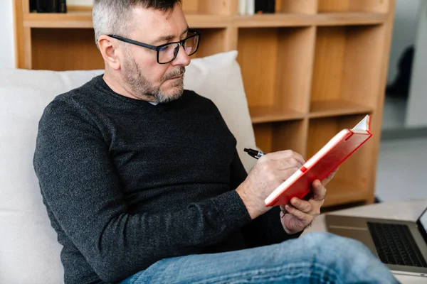 Ernsthafter Erwachsener Unrasierter Mann Schreibt Notizen Während Hause Mit Laptop — Stockfoto